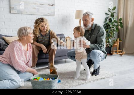 Ein fröhlicher bärtiger Mann, der ein kleines Mädchen unterstützt, der seine ersten Schritte in der Nähe der Familie im Wohnzimmer macht, Stockbild Stockfoto