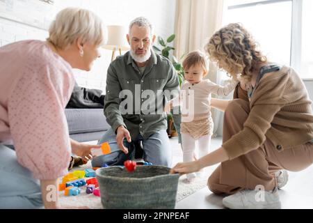 Blonde Frau mit Kleinkindertochter und reife Eltern in der Nähe von Korb und Spielzeug auf dem Boden im Wohnzimmer, Stockbild Stockfoto