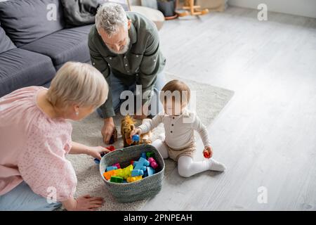 Großer Blickwinkel auf ältere Paare und Kleinkinder in der Nähe des Korbs mit Spielzeug auf dem Boden im Wohnzimmer, Stockbild Stockfoto