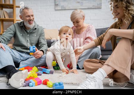 Ein kleines Mädchen, das in der Nähe von Spielzeugen wegschaut und eine glückliche Familie auf dem Boden im Wohnzimmer hat, Stockbild Stockfoto