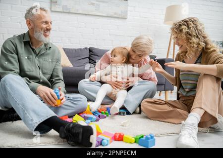 Reife Frau umarmt Enkeltochter in der Nähe von Spielzeug und fröhliche Familie auf dem Boden im Wohnzimmer, Stockbild Stockfoto