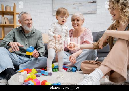 Fröhliche Frau, die die Hand der kleinen Tochter in der Nähe glücklicher Eltern hält und Spielzeug auf dem Boden im Wohnzimmer, Stockbild Stockfoto
