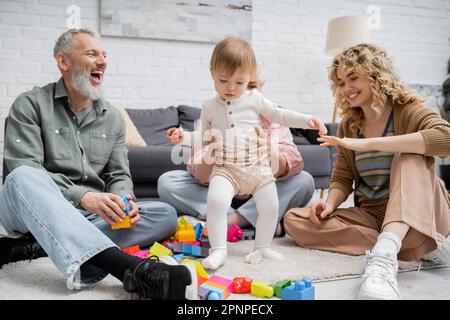 Aufgeregter bärtiger Mann, der in der Nähe einer Familie lacht und mit einem Kind auf dem Boden im Wohnzimmer spielt, Stockbild Stockfoto