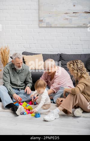 Glückliches Paar mittleren Alters, das Bausteine spielt, mit der Enkelin auf dem Boden im Wohnzimmer, Stockbild Stockfoto