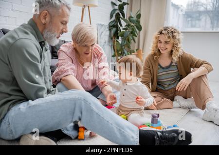 Fröhliche Frau, die Eltern mittleren Alters ansieht, die Bausteine spielen, mit Kind auf dem Boden im Wohnzimmer, Bild der Aktien Stockfoto