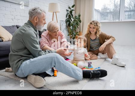 Fröhliches, reifes Paar, das Bausteine mit seiner Enkelin spielt, während es auf dem Boden im modernen Wohnzimmer sitzt, Stockbild Stockfoto