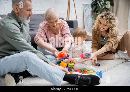 Kleinkindermädchen spielt mit bunten Bausteinen in der Nähe von glücklichen Eltern und Großeltern auf dem Boden im Wohnzimmer, Stockbild Stockfoto