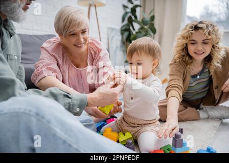 Lächelnde Großeltern geben Kleinkindern Spielzeug, während sie im Wohnzimmer spielen, Stockbild Stockfoto