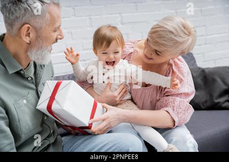 Sorgenfreies Baby lacht und Gesten in der Nähe lächelnder Großeltern und präsent auf dem Sofa zu Hause, Stockbild Stockfoto