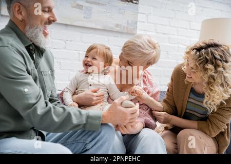 Ein bärtiger Mann berührt das Bein einer überfreulichen Enkelin, während er mit der Familie auf der Couch im Wohnzimmer Spaß hat, Stockbild Stockfoto