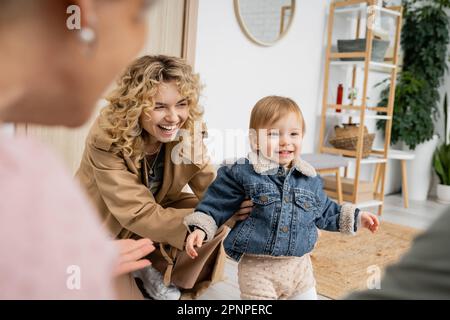 Fröhliches Kind in Jeansjacke lächelnd neben glücklicher Mutter und Oma auf verschwommenem Vordergrund, Stockbild Stockfoto