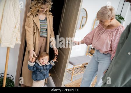 Glückliche Frau mittleren Alters öffnet die Tür zur Tochter mit Enkel in Jeansjacke während ihres Besuchs zu Hause, Stock Image Stockfoto