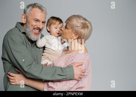 Reife Frau küsst Enkeltochter nahe eines glücklichen bärtigen Ehemanns isoliert auf grauem, Stock-Bild Stockfoto