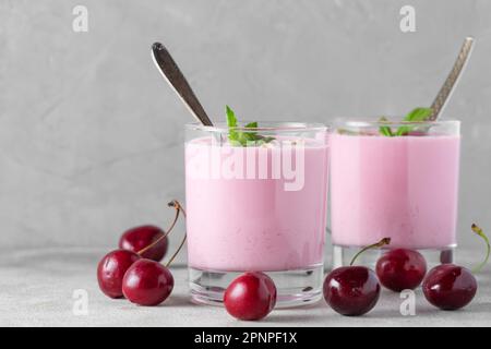 Zwei Gläser gesunder Kirschjoghurt mit frischen Beeren, Hafer, Löffel und Minze auf grauem Hintergrund. Gesundes Frühstück. Leckeres Essen Stockfoto
