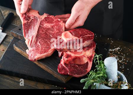 Steak-Rindfleisch. Der Koch schneidet Steak-Rindfleisch. Die Hände von Mans halten rohes Tomahawk-Steak auf rustikalem Holzbrett auf schwarzem Hintergrund. Kochen, Rezepte und Stockfoto