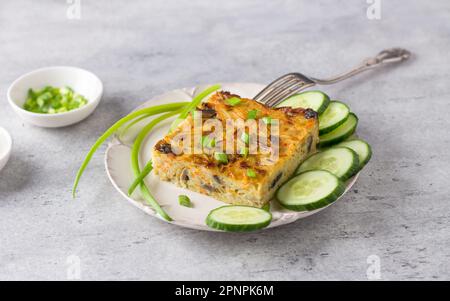 Gemüseauflauf, kugel, aus Kartoffeln, Karotten und Pilzen mit Gurkenscheiben und grünen Zwiebeln auf grauem, strukturiertem Hintergrund. Köstliche Vegeta Stockfoto