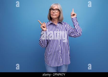 Überraschte Frau mittleren Alters mit grauen Haaren zeigt mit dem Finger zur Seite auf einem hellen Studiohintergrund mit Kopierbereich Stockfoto
