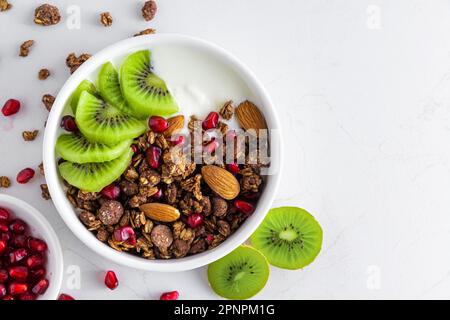 Griechischer Joghurt mit Schokoladengranola, Kiwis, Granatapfelsamen und Mandeln in einer Schüssel für ein gesundes Frühstück auf weißem Hintergrund. Draufsicht Stockfoto