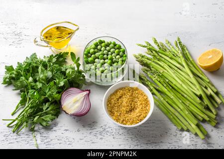 Zutaten für einen Tabbouleh-Salat aus dem Nahen Osten. Bulgur, frischer junger grüner Spargel, grüne Erbsen, Tomaten, rote Zwiebeln, Kräuter, Zitrone und Olivenöl auf einem Stockfoto