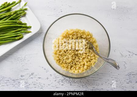Glasschüssel mit Bulgur und einem Teller mit Spargel auf hellblauem Hintergrund, Draufsicht. Köstlicher Hausgemachter Vegan-Spargel Tabbouleh-Salat Stockfoto