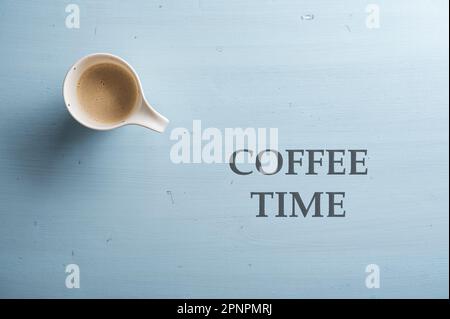 Draufsicht einer Tasse frischen Kaffees auf einem einfachen hellblauen Holzhintergrund. Neben der Kaffeezeit steht ein Schild mit schwarzem Großbuchstaben Stockfoto