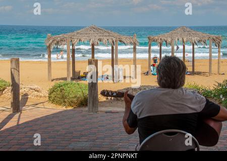 Ein Mann mit Gitarre sitzt mittags und spielt im Schatten am Strand Stockfoto