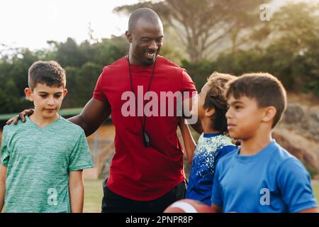 Fröhlicher Trainer mit seinem Rugby-Team der Grundschule. Sportlehrer, der eine Gruppe von Kindern in der Grundschule ausbildet. Mentorenstudium und D Stockfoto