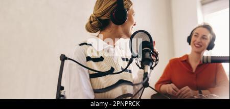 Fröhlicher Radio-Gastgeber, der einen Gast in ihrer Show in ihrem Heimstudio interviewt. Zwei fröhliche Frauen, die sich amüsieren, während sie eine Live-Audioübertragung auf aufnehmen Stockfoto