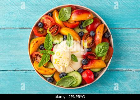 Köstlicher Sommersalat mit Burrata-Käse, gegrillten Pfirsichen, Tomaten, Heidelbeeren, Gurken, Olivenöl und Basilikum. Draufsicht. Gesunde Ernährung Stockfoto