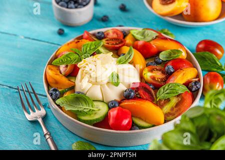 Köstlicher Sommersalat mit Burrata-Käse, gegrillten Pfirsichen, Tomaten, Blaubeeren, Gurke und Basilikum auf einem blauen Tisch. Gesunde vegetarische Ernährung Stockfoto