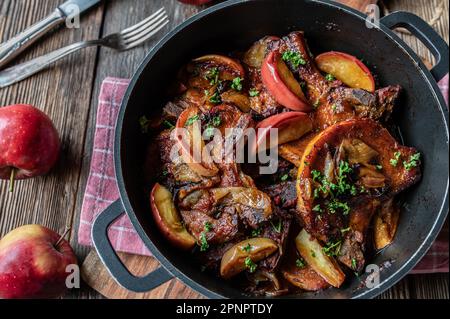 Geschmorte Schweinekoteletts mit Äpfeln und Zwiebeln in einem Brattopf auf einem Holztisch Stockfoto