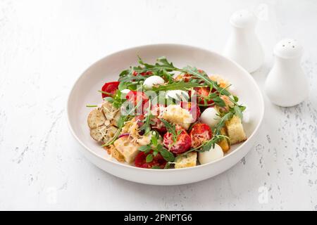Gebackene Panzanella, traditioneller italienischer Salat mit Tomaten, Mozzarella, Parmesan, knusprigen Croutons, Rucola und Basilikum auf hellblauem Hintergrund Stockfoto