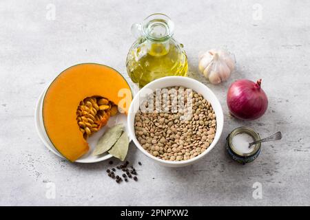 Zutaten zum Kochen von köstlichem veganem Eintopf: Kürbis, grüne Linsen, rote Zwiebel, Knoblauch, Olivenöl, Salz, schwarzer Pfeffer, Lorbeerblatt auf einem grauen, strukturierten Ba Stockfoto