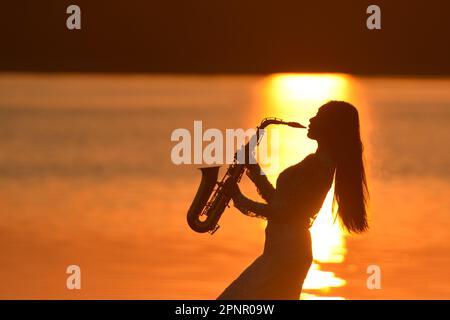 Silhouette einer weiblichen Saxophonistin, die das Saxophon bei Sonnenuntergang am See spielt, Thailand Stockfoto