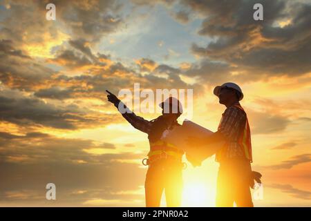 Silhouette von zwei Bauingenieuren, die auf einer Baustelle stehen und ein Bauprojekt bei Sonnenuntergang in Thailand beaufsichtigen Stockfoto