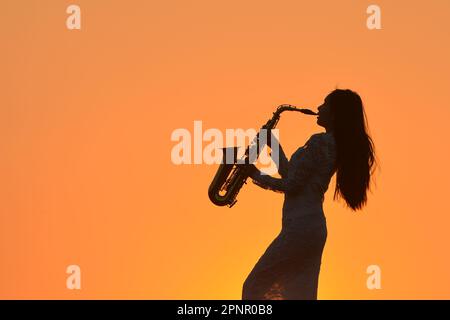 Silhouette einer weiblichen Saxophonistin, die das Saxophon bei Sonnenuntergang am See spielt, Thailand Stockfoto