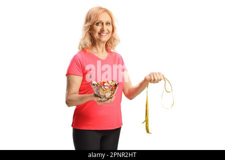 Schöne, reife Frau in Sportbekleidung, die einen Salat und ein Maßband auf weißem Hintergrund hält Stockfoto