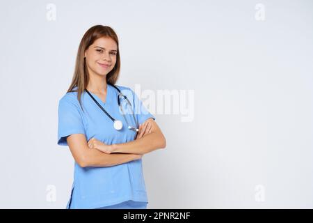 Porträt einer lächelnden Ärztin im blauen Mantel, die mit gefalteten Armen auf weißem Hintergrund posiert. Stockfoto