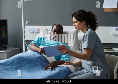 Junge Krankenschwester zeigt ältere Patienten auf einem digitalen Tablet ihre Krankengeschichte, während sie auf der Station im Bett liegt Stockfoto