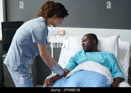 Die junge Krankenschwester, die Tropfer für kranke Patienten herstellt, führte einen Katheter in seinen Arm ein, während er auf dem Bett auf der Station lag Stockfoto