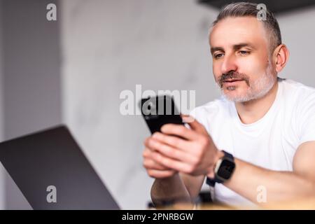 Ein alter Mann sitzt in der Küche neben einem Laptop und hält ein Smartphone mit einem Gerät, chattet aus der Ferne, durchblättert die Webseite, nutzt die neue mobile App Genießen Sie die Nutzung der modernen Wireless-Technologie, ma Stockfoto