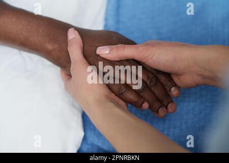 Nahaufnahme einer jungen Krankenschwester, die mit einem älteren afroamerikanischen Mann Händchen hält und ihn während seiner Krankheit unterstützt Stockfoto