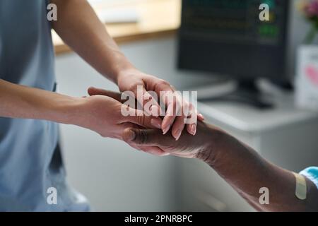Nahaufnahme einer jungen Ärztin, die Händchen mit einem afroamerikanischen Patienten hält, während er auf der Station liegt Stockfoto