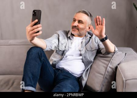 Seitenansicht eines glücklichen, gutaussehenden Mannes, der über das Smartphone eine Videokonferenz mit Freunden oder Liebhabern hat, sich im Wohnzimmer auf der Couch zurücklehnen und beim mobilen Telefon winken kann Stockfoto