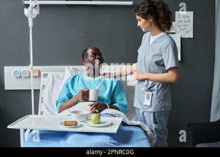 Junge Krankenschwester in Uniform serviert dem Seniorpatienten Mittagessen, während er auf der Station im Bett sitzt Stockfoto