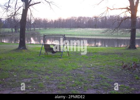 Eine getarnte Schaukel befindet sich im Hof in der Nähe eines Teichs, ein entspannender Ort zum Sitzen und Genießen des Tages. Stockfoto