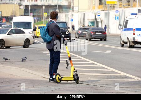 Ein Mann mit Kopfhörern, der mit einem elektrischen Roller in der Frühlingsstadt auf Autos steht Stockfoto