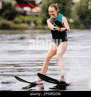 Lächelnde junge Frau, Wasserski hinter dem Motorboot auf dem Teich, schöne Sommerferien Stockfoto