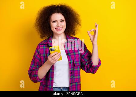 Foto des stylischen jungen Damen-Haarstils mit OKY Symbol Hold Smartphone empfehlen Internetshop-Angebote isoliert auf gelbem Hintergrund Stockfoto