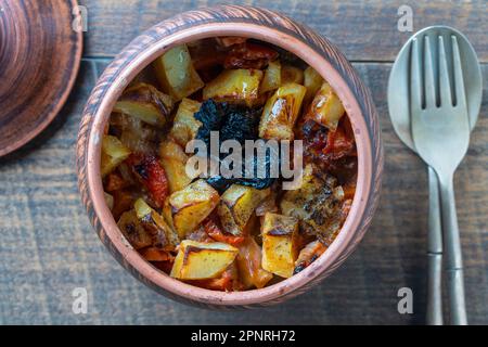 Gedünstete Kartoffeln, Karotten, Zwiebeln, Tomaten und Backpflaumen in einem Tontopf auf Holztisch-Hintergrund, Nahaufnahme, Draufsicht Stockfoto